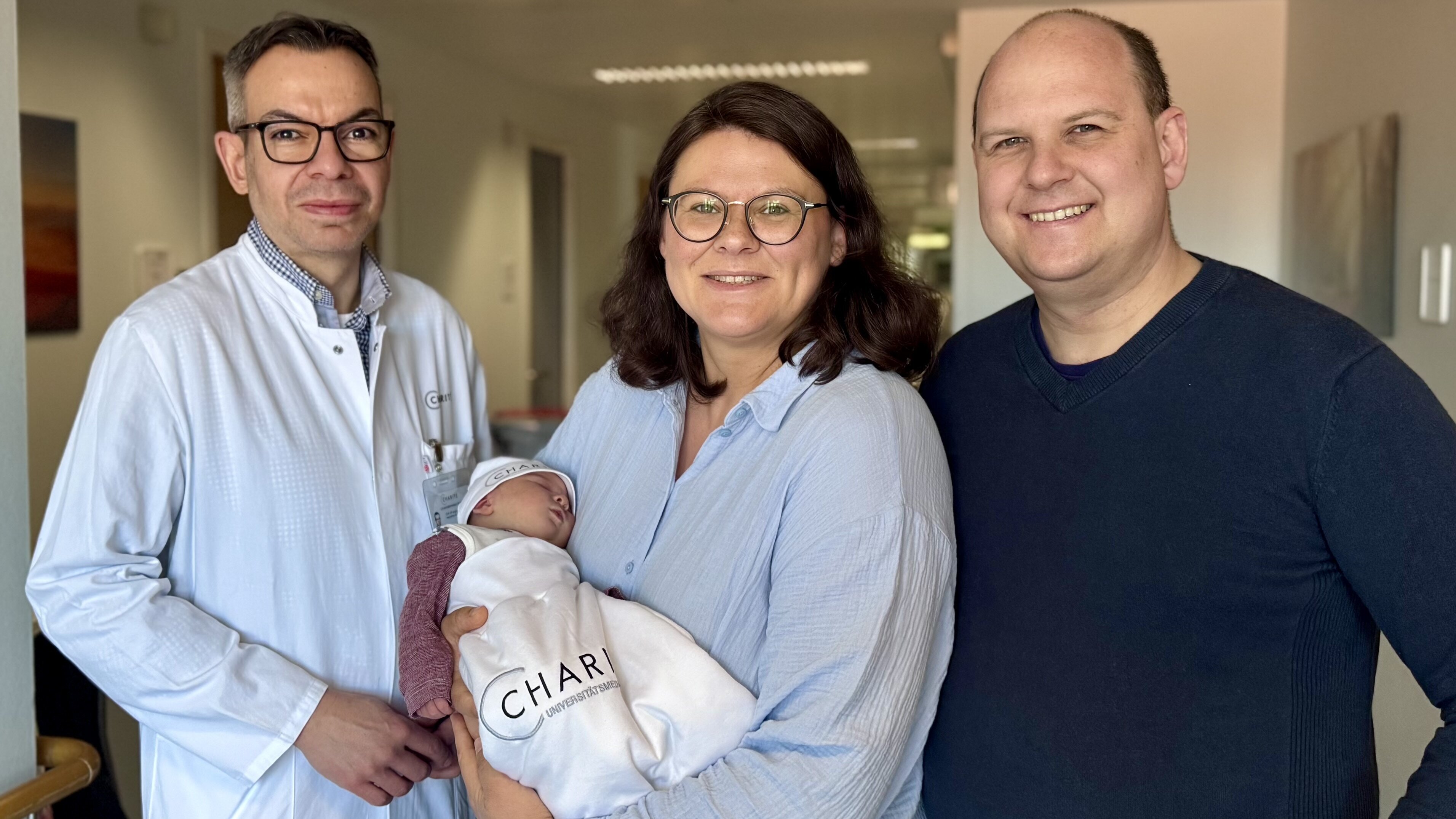 Ein Arzt in weißem Kittel steht neben einer lächelnden Frau, die ein Baby in einer Decke hält. Neben ihnen steht ein lächelnder Mann. Die Atmosphäre ist freundlich und feierlich, was auf die Geburt des Babys hindeutet.