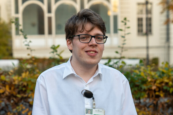 Ein junger Mann mit Brille und einem weißen Hemd lächelt in die Kamera. Er trägt einen Ausweis am Hals und steht vor einer grünen Hecke mit gelben Blättern im Hintergrund. Die Szene vermittelt eine freundliche und einladende Atmosphäre.