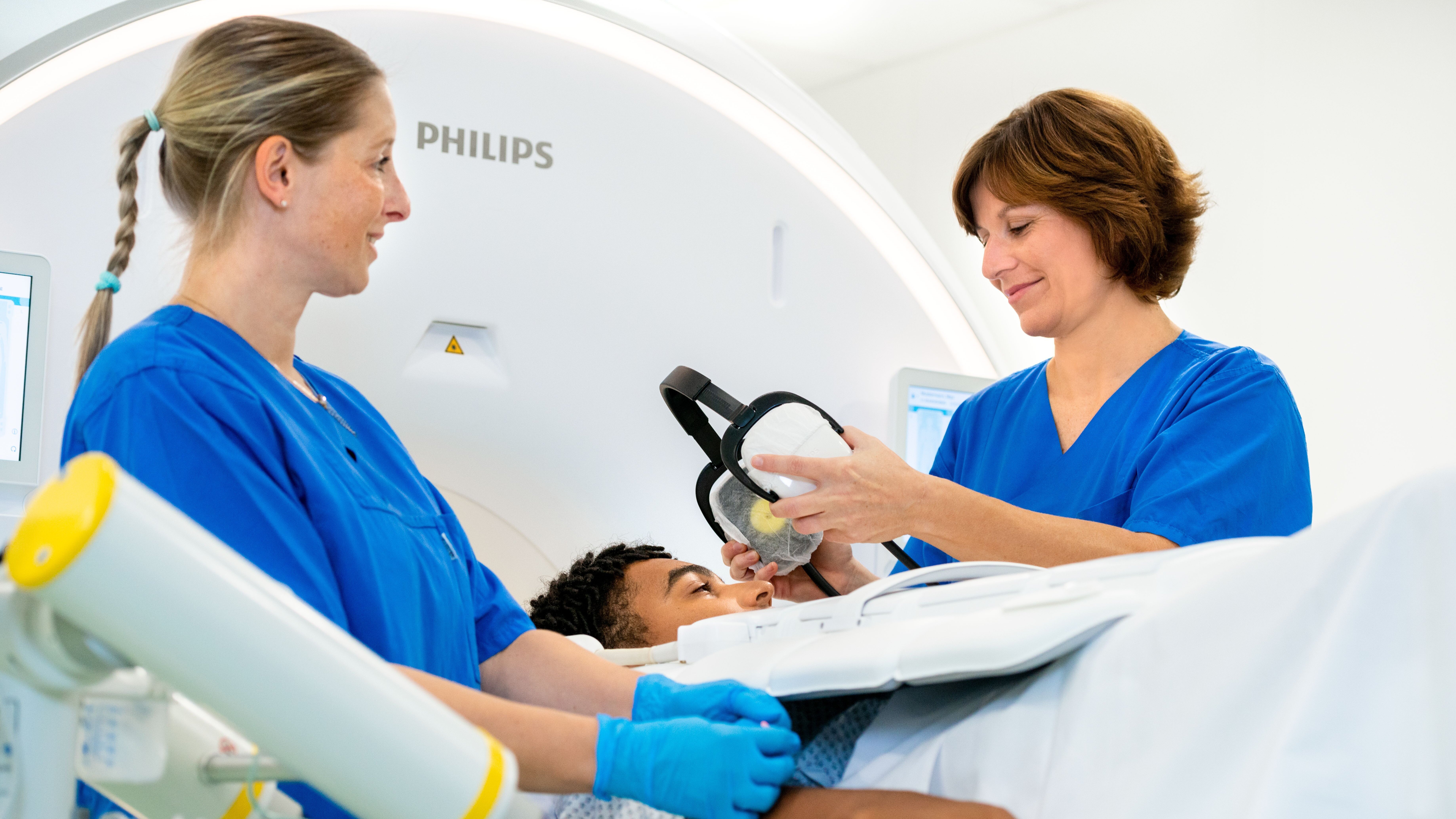 Zwei medizinische Fachkräfte in blauen Kitteln stehen neben einem Patienten, der auf einer Liege liegt und sich einer Untersuchung unterzieht. Eine der Fachkräfte hält eine Maske in der Hand, während die andere den Patienten beruhigend anfasst. Die Ausstattung deutet auf eine medizinische Umgebung hin.