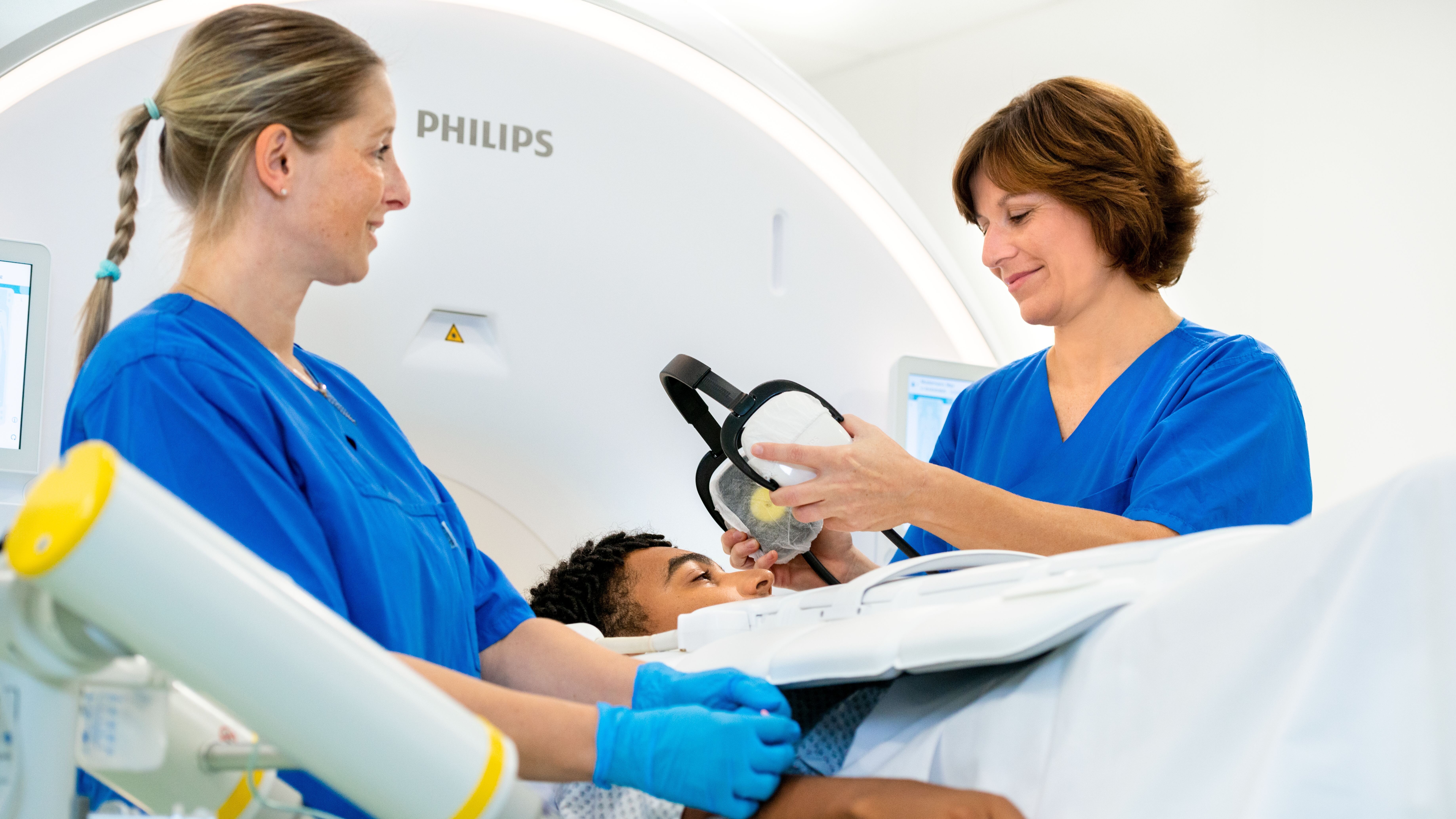 Zwei medizinische Fachkräfte in blauen Kitteln stehen neben einem Patienten, der auf einer Liege liegt und sich einer Untersuchung unterzieht. Eine der Fachkräfte hält eine Maske in der Hand, während die andere den Patienten beruhigend anfasst. Die Ausstattung deutet auf eine medizinische Umgebung hin.