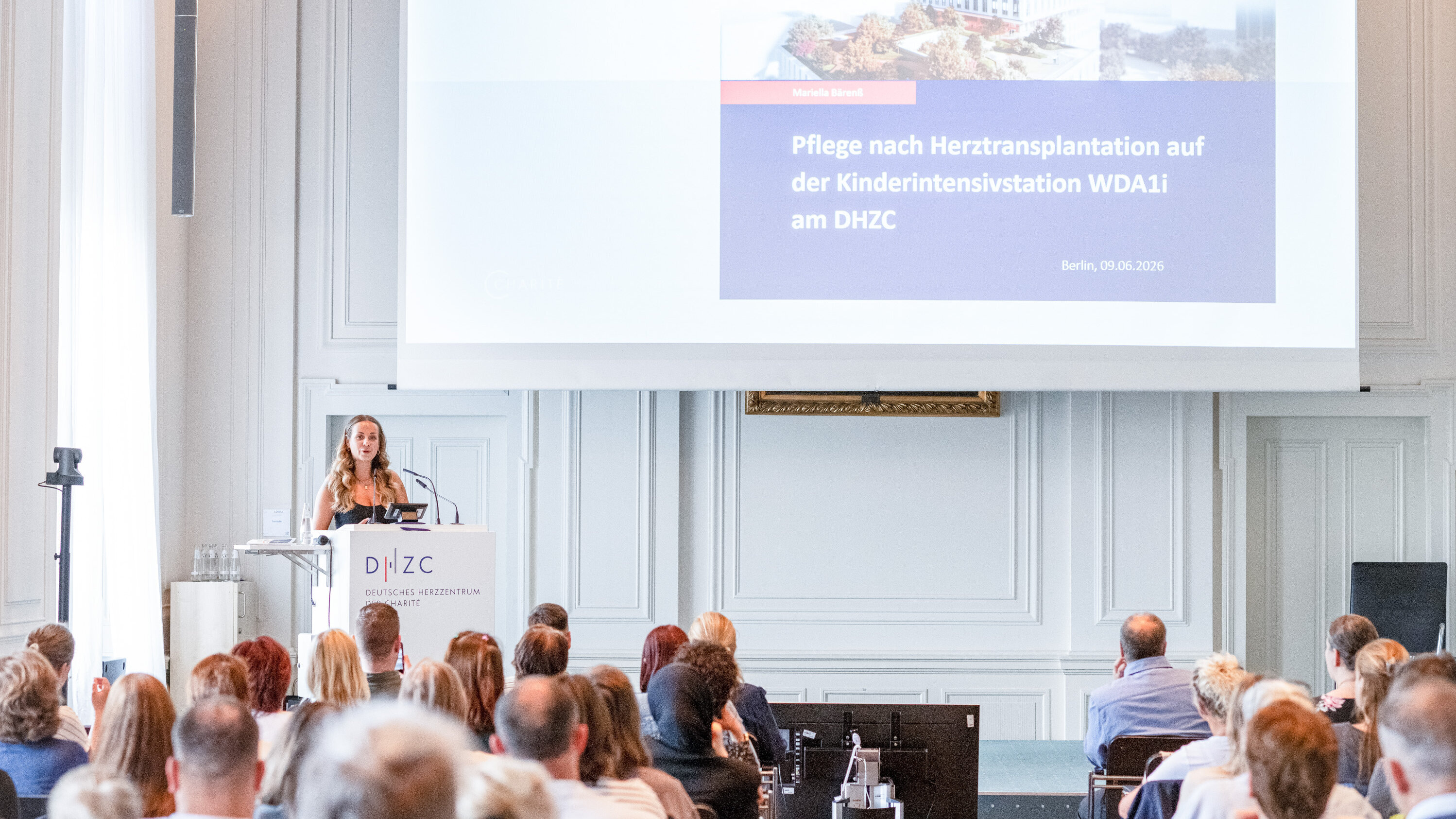 Eine Präsentatorin steht an einem Rednerpult vor einer großen Gruppe von Menschen. Im Hintergrund wird eine Folie mit dem Titel "Pflege nach Herztransplantation auf der Kinderintensivstation WDA1 am DHZC" angezeigt. Der Raum ist hell und modern eingerichtet.