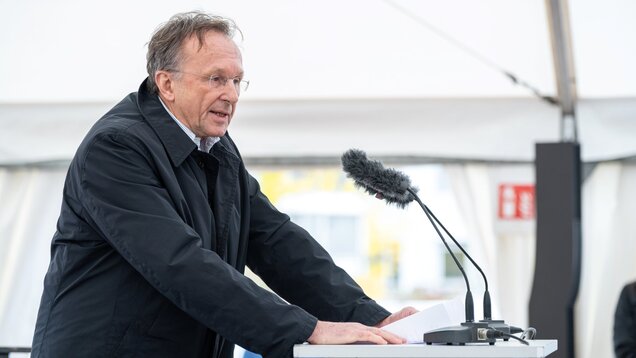 Ein Mann spricht am Rednerpult unter einem Zelt. Er trägt eine schwarze Jacke und hat eine Notiz in der Hand. Ein Mikrofon ist am Pult angebracht. Im Hintergrund sind verschwommene Personen und eine helle Umgebung zu erkennen.