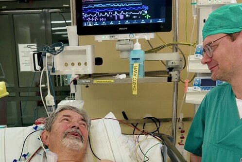 Ein älterer Mann liegt im Krankenhausbett und schaut lächelnd einen Arzt an, der neben ihm steht. Der Mann ist mit einem weißen Laken und medizinischen Leitungen bedeckt. Im Hintergrund sind Monitore mit Vitalzeichenanzeigen zu sehen.