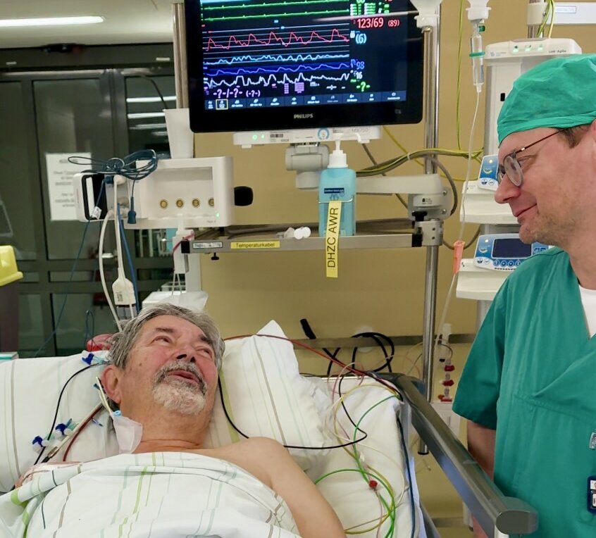 Ein älterer Mann liegt im Krankenhausbett und schaut lächelnd einen Arzt an, der neben ihm steht. Der Mann ist mit einem weißen Laken und medizinischen Leitungen bedeckt. Im Hintergrund sind Monitore mit Vitalzeichenanzeigen zu sehen.