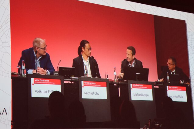 Eine Diskussionsrunde mit vier Teilnehmern auf einer Bühne. Die Referenten, namentlich Volkmar Fallak, Michael Chu, Michael Berger und Antonios Pitsik, sitzen an einem Tisch und diskutieren vor einem roten Hintergrund. Es sind Mikrofone und Laptops sichtbar.