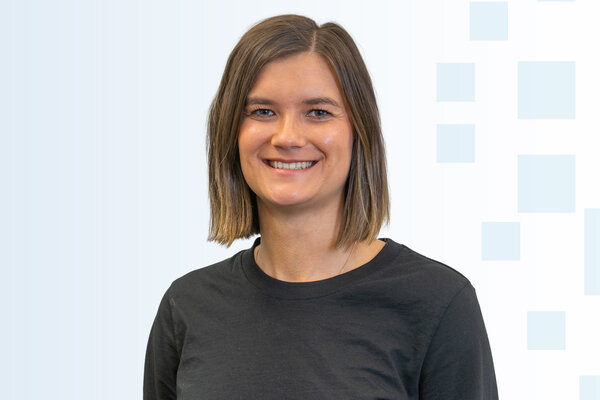 Ein lächelnde Frau mit schulterlangen, braunen Haaren trägt ein schwarzes T-Shirt. Der Hintergrund ist hell mit blauen Quadraten.