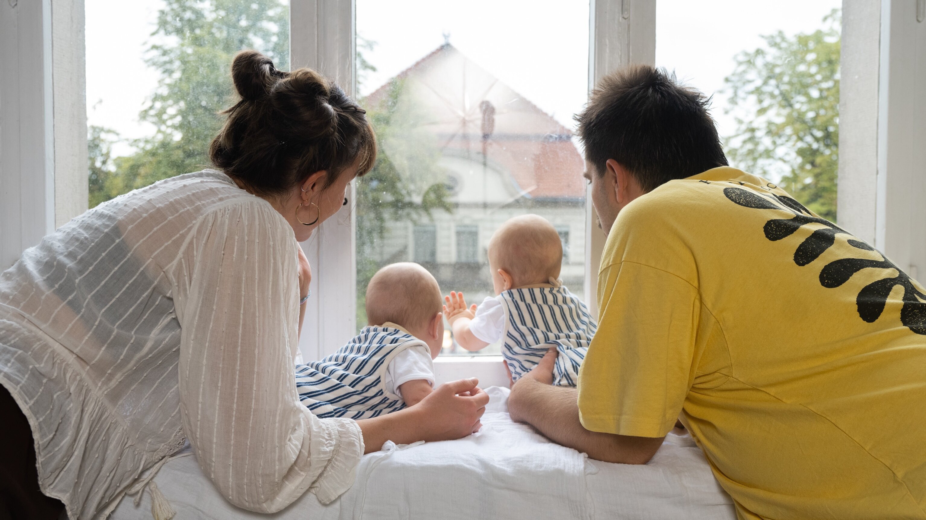 Eine Frau und ein Mann lehnen sich über eine Fensterbank, während zwei Babys in Streifenoveralls nach draußen schauen. Die Szene vermittelt Neugier und Familienzeit in einem hellen Raum mit Blick auf einen Garten.