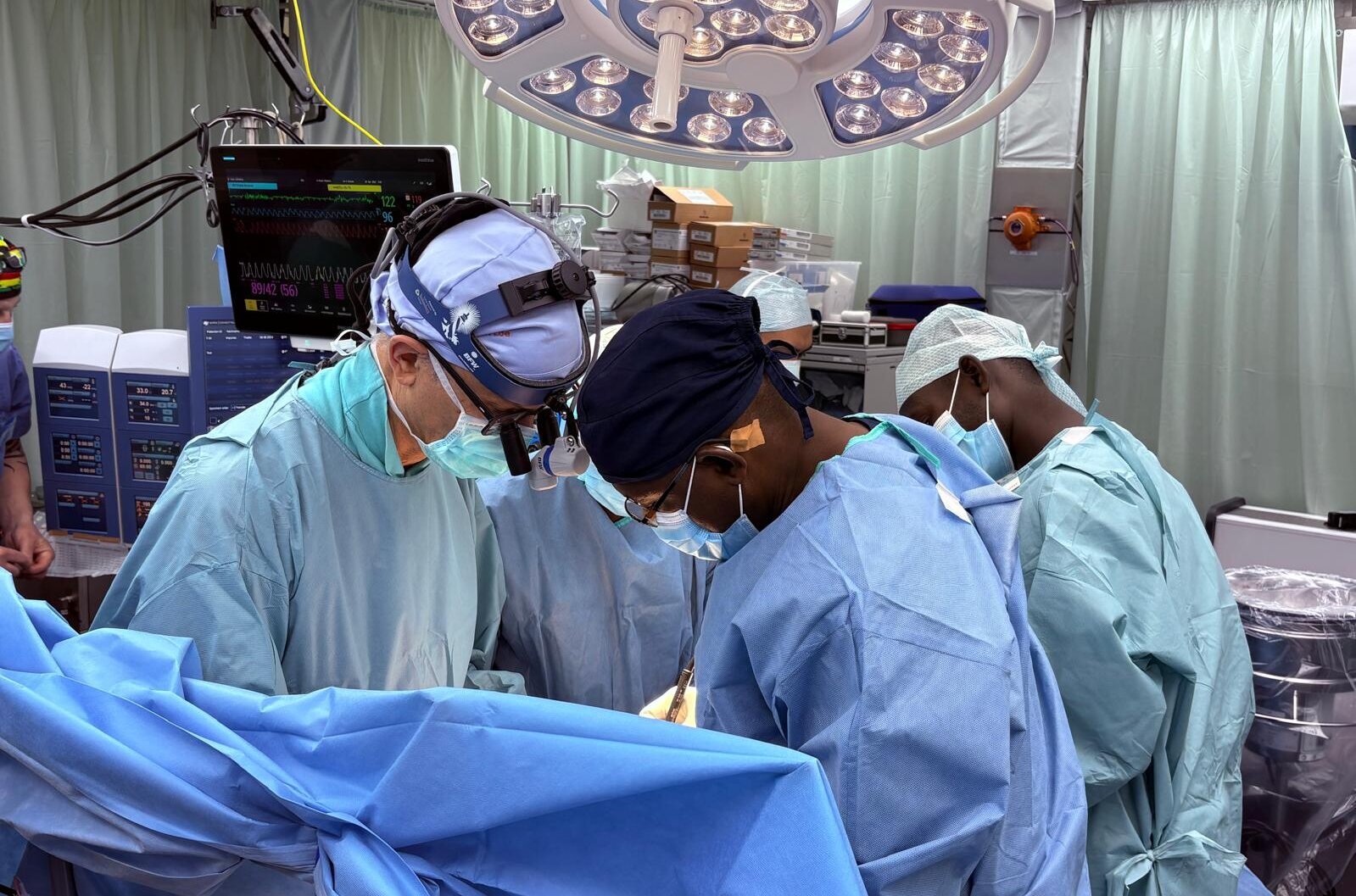 Vier Chirurgen in Operationskleidung arbeiten konzentriert an einem chirurgischen Eingriff. Sie tragen Masken und Hauben, während sie über einem Patienten stehen, der von blauen Tüchern bedeckt ist. Um sie herum befinden sich medizinische Geräte und Beleuchtung.