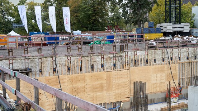 Eine Baustelle mit einem tiefen Loch in der Erde, umgeben von Holzstützen und Unterstützungselementen. Im Hintergrund sind Baugeräte und Materialien, einschließlich grüner Rohre und Beton, sichtbar. Die Umgebung besteht aus Bäumen und Baustellenmaterial.