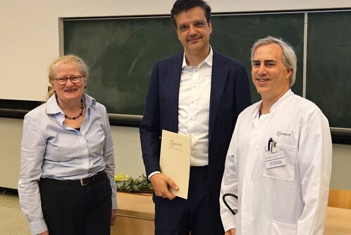 Drei Personen stehen in einem Raum. Links ist eine Frau mit Brille und blauer Bluse. In der Mitte steht ein Mann in einem dunklen Anzug, der ein Dokument hält. Rechts steht ein Mann in einem weißen Kittel mit grünen Schuhen. Im Hintergrund ist eine Tafel sichtbar.