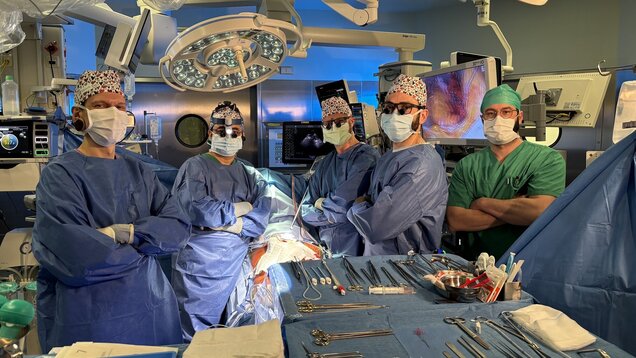Fünf medizinische Fachkräfte stehen in einem Operationssaal. Sie tragen OP-Kleidung, Masken und Haarnetze. In der Mitte des Raumes sind chirurgische Instrumente und ein OP-Tisch zu sehen. Der Raum ist gut beleuchtet und der Hintergrund zeigt medizinische Geräte.
