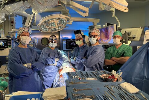 Fünf medizinische Fachkräfte stehen in einem Operationssaal. Sie tragen OP-Kleidung, Masken und Haarnetze. In der Mitte des Raumes sind chirurgische Instrumente und ein OP-Tisch zu sehen. Der Raum ist gut beleuchtet und der Hintergrund zeigt medizinische Geräte.