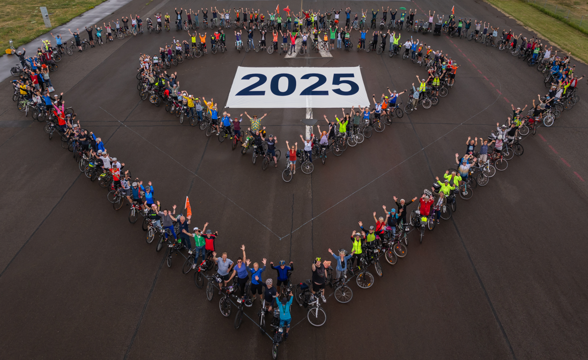 Eine Gruppe von Radfahrern bildet in einer herzförmigen Anordnung die Zahl "2025" auf einem Asphaltplatz. Die Teilnehmer tragen verschiedene bunte Radkleidung und heben die Arme zur Feier. Die Szene vermittelt Gemeinschaft und Vorfreude auf das Jahr 2025.