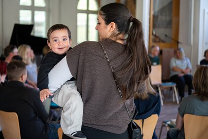 Eine Frau hält ein lächelndes Kleinkind auf dem Arm. Im Hintergrund sind mehrere Erwachsene in einer Umgebungsstruktur zu sehen, die sich in einem Raum befinden. Das Bild vermittelt eine freundliche und familiäre Atmosphäre.