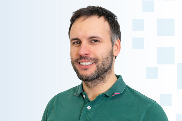 Ein junger Mann mit Bart lächelt freundlich in die Kamera. Er trägt ein grünes Polohemd und hat kurze Haare. Der Hintergrund besteht aus hellen Farben und geometrischen Formen.