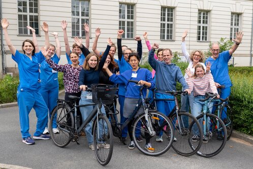 Eine Gruppe von Menschen in blauen und bunten Outfits steht auf einem Gehweg und hebt die Hände. Einige Personen halten Fahrräder. Die Atmosphäre ist fröhlich und einladend, was auf ein gemeinschaftliches oder feierliches Ereignis hinweist.