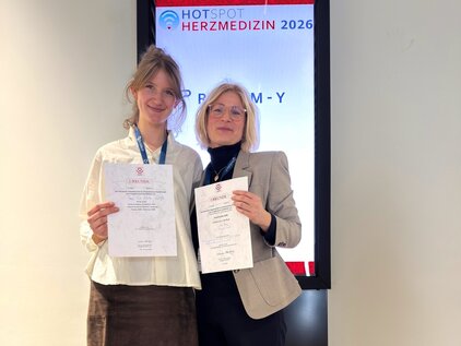 Zwei Frauen stehen nebeneinander und halten jeweils ein Dokument in der Hand. Im Hintergrund befindet sich ein Bildschirm mit der Aufschrift „HOTSPOT HERZMEDIZIN 2026“. Beide Frauen tragen formelle Kleidung und lächeln, was auf einen feierlichen Anlass hinweist.