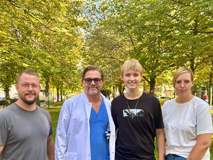 Vier Personen stehen unter Bäumen in einem Park. Eine Person trägt einen weißen Kittel und zeigt eine freundliche Haltung. Die anderen drei sind lässig gekleidet und lächeln. Die Szene vermittelt eine positive, gemeinschaftliche Atmosphäre.