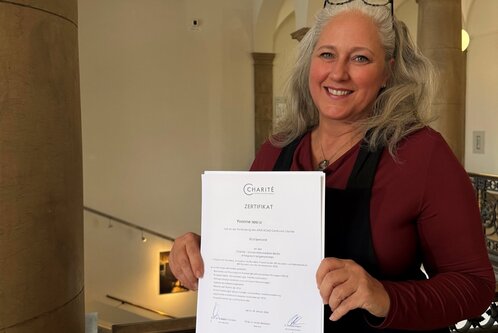 Eine lächelnde Frau mit langen, lockigen Haaren hält stolz ein Zertifikat in der Hand. Sie trägt ein schwarzes Oberteil und steht in einem historischen Gebäude mit einer eleganten Treppe im Hintergrund.