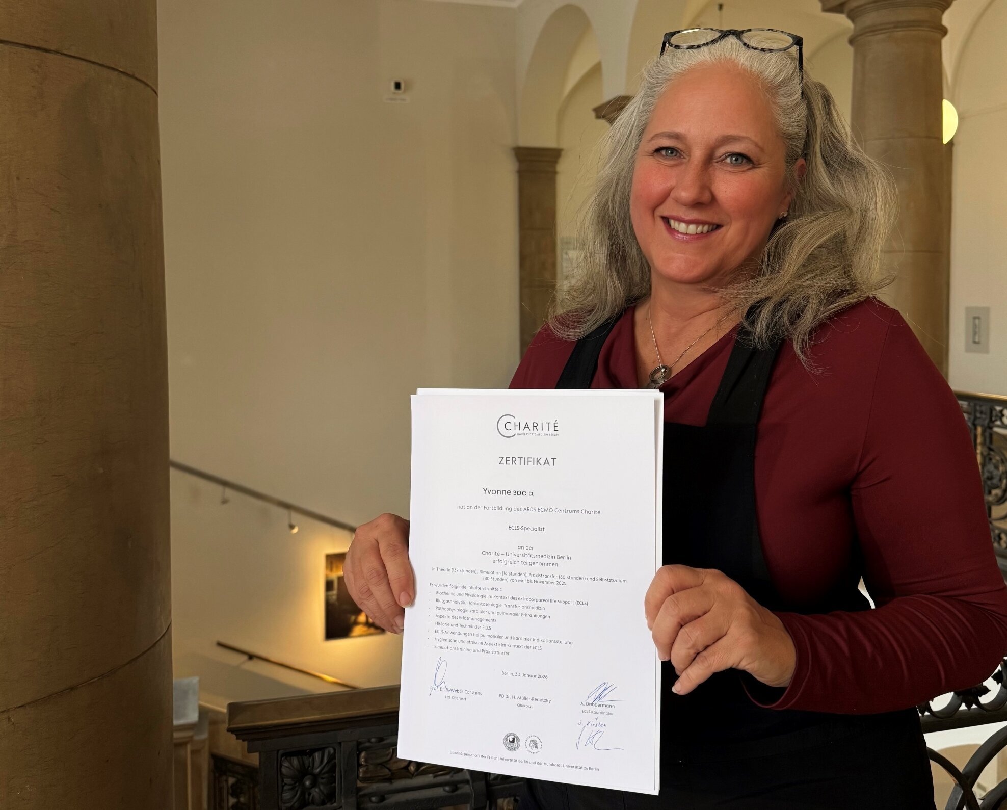 Eine lächelnde Frau mit langen, lockigen Haaren hält stolz ein Zertifikat in der Hand. Sie trägt ein schwarzes Oberteil und steht in einem historischen Gebäude mit einer eleganten Treppe im Hintergrund.