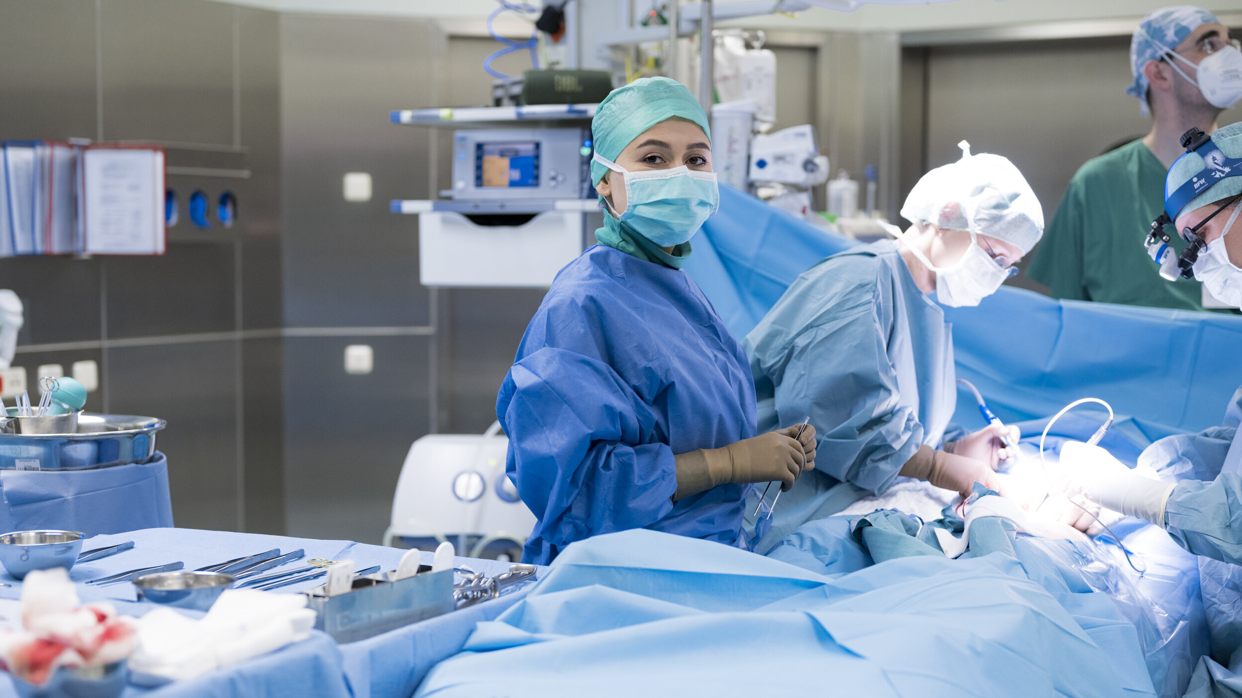 Ein Operationsteam in einem modernen Operationssaal führt einen Eingriff durch. Ärzte in blauen OP-Kitteln und Schutzmasken konzentrieren sich auf die Operation. Instrumente und sterile Materialien sind auf dem Tisch in der Nähe angeordnet.