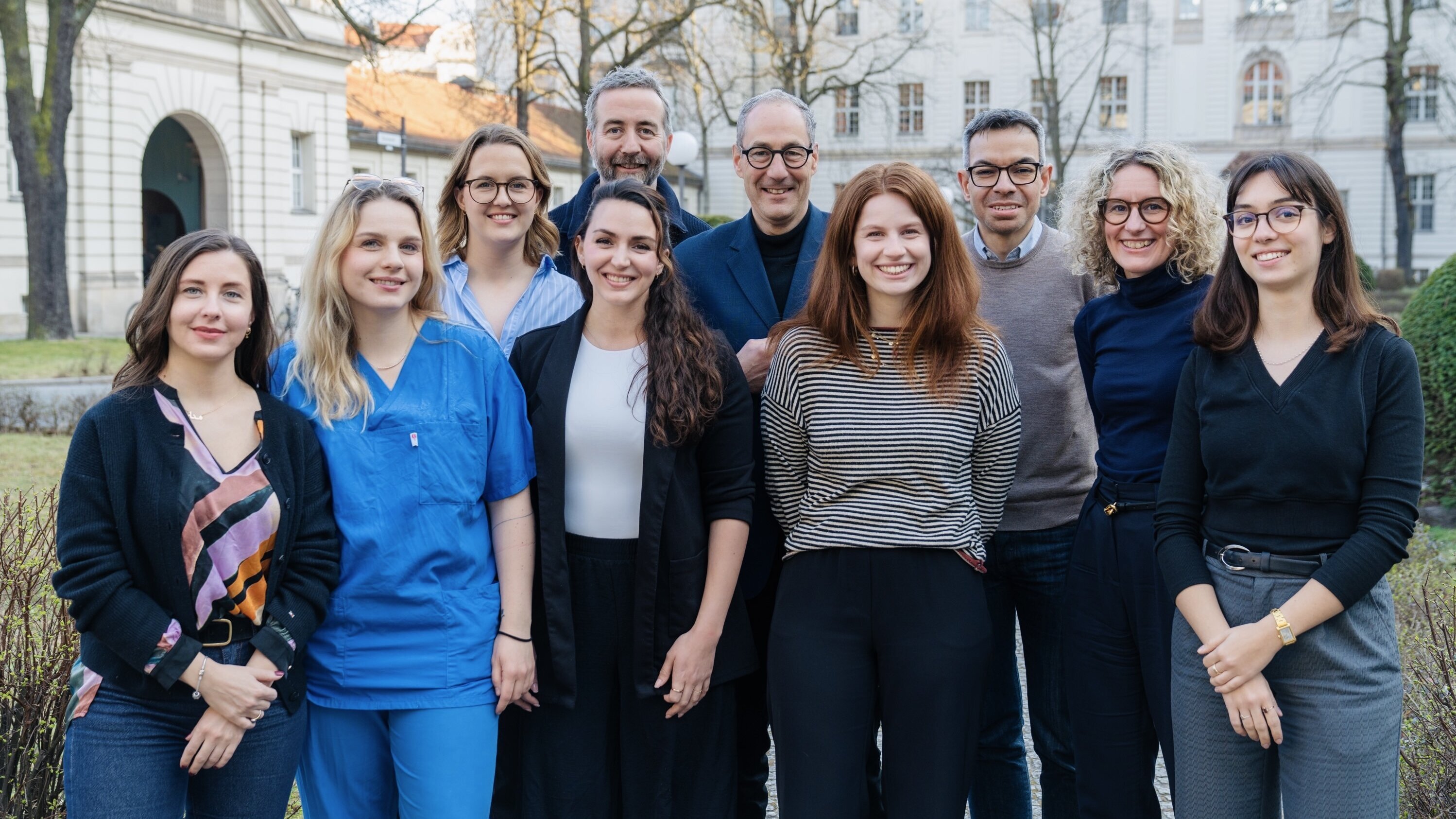 Eine Gruppe von zehn Personen steht im Freien vor einem historischen Gebäude. Sie lächeln und posieren gemeinsam. Die Bildunterschrift könnte darauf hinweisen, dass es sich um ein Team oder eine Gemeinschaft handelt. Der Hintergrund zeigt Bäume und einen gepflegten Garten.