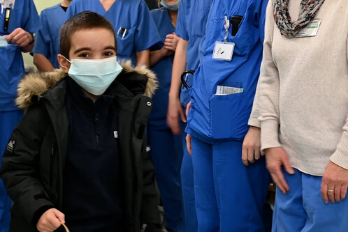 Ein lächelnder Junge mit einem Mundschutz trägt eine Jacke mit Fellkragen. Um ihn herum stehen mehrere medizinische Fachkräfte in blauen Kitteln. Die Gruppe wirkt freundlich und unterstützend. Der Junge hält einen Stock in der Hand, während er in die Kamera schaut.