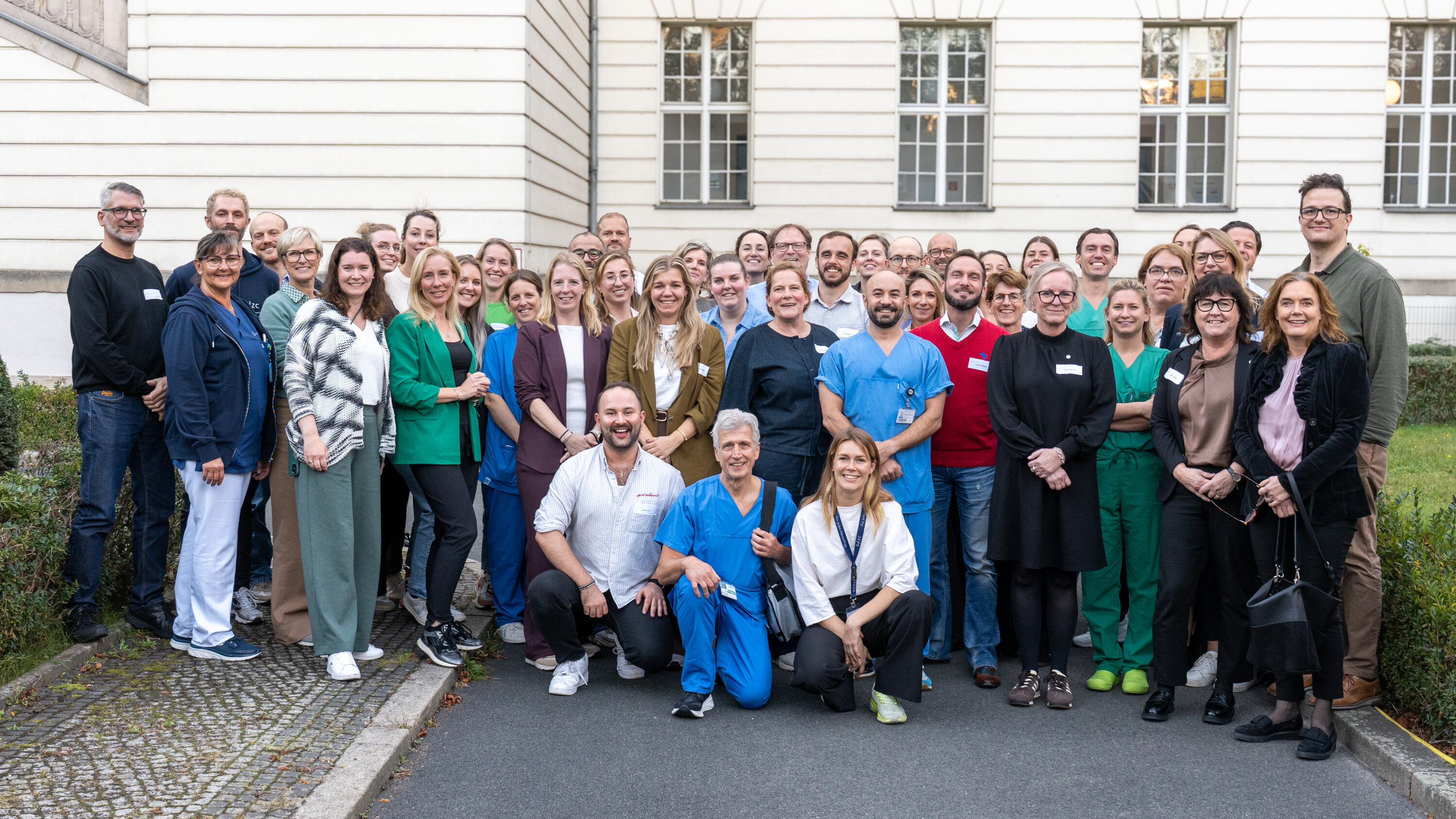 Eine große Gruppe von Menschen steht auf einem Hof vor einem Gebäude. Die Teilnehmer tragen unterschiedliche Arbeitskleidung, darunter medizinische Uniformen. Sie lächeln und posieren für das Foto, darunter Ärzte, Pflegekräfte und weitere Fachkräfte.