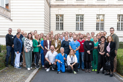 Eine große Gruppe von Menschen steht auf einem Hof vor einem Gebäude. Die Teilnehmer tragen unterschiedliche Arbeitskleidung, darunter medizinische Uniformen. Sie lächeln und posieren für das Foto, darunter Ärzte, Pflegekräfte und weitere Fachkräfte.