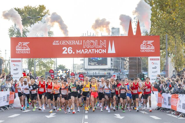 Start des 26. Generali Köln Marathons: Eine Gruppe von Läufern steht an der Startlinie, während eine große Banner mit dem Schriftzug „Köln Marathon“ im Hintergrund prangt. Rauch geht von Pyrotechnik aus, und Zuschauer sind an der Strecke versammelt.