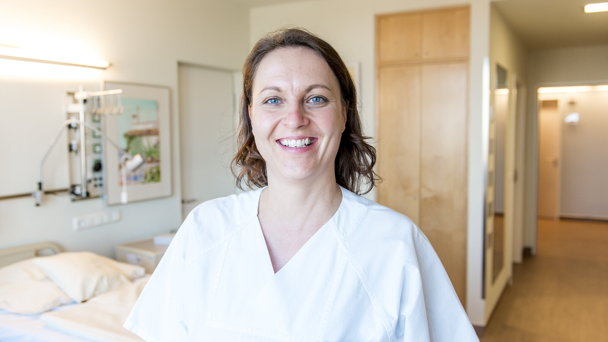 Eine lächelnde Pflegekraft in weißer Uniform steht in einem modernen Krankenhauszimmer. Im Hintergrund sind ein Bett und medizinische Geräte sichtbar. Die Person strahlt Wärme und Freundlichkeit aus.