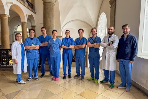 Eine Gruppe von acht medizinischen Fachkräften in blauen Uniformen steht lächelnd in einem Innenbereich mit hohen Decken und großen Fenstern. Sie zeigen eine freundliche Haltung und scheinen zusammenzuarbeiten. Ein Mitglied trägt einen weißen Kittel, während die anderen in blauen Scrubs gekleidet sind.
