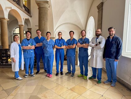 Eine Gruppe von acht medizinischen Fachkräften in blauen Uniformen steht lächelnd in einem Innenbereich mit hohen Decken und großen Fenstern. Sie zeigen eine freundliche Haltung und scheinen zusammenzuarbeiten. Ein Mitglied trägt einen weißen Kittel, während die anderen in blauen Scrubs gekleidet sind.