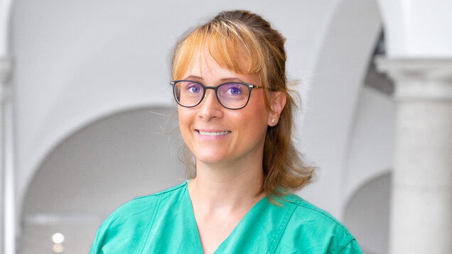 Eine lächelnde Frau mit blonden, halblangen Haaren trägt ein grünes medizinisches Outfit und Brille. Sie steht vor einer hellen, architektonischen Kulisse mit Bögen und Säulen. Ihr Blick ist freundlich und einladend.