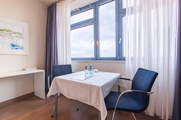 Ein heller Raum mit großen Fenstern, durch die Tageslicht strömt. In der Mitte steht ein Tisch mit einer weißen Tischdecke, auf dem zwei Flaschen Wasser und ein Glas stehen. Zwei blaue Stühle sind um den Tisch platziert.
