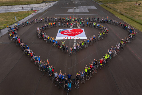 Eine Gruppe von Radfahrern steht auf einem Flugplatz und bildet gemeinsam ein Herz. In der Mitte liegt ein Banner mit der Aufschrift "PSG HereFahrt 2025". Viele Teilnehmer heben die Hände und sind in bunten Outfits gekleidet. Es herrscht eine fröhliche Atmosphäre.