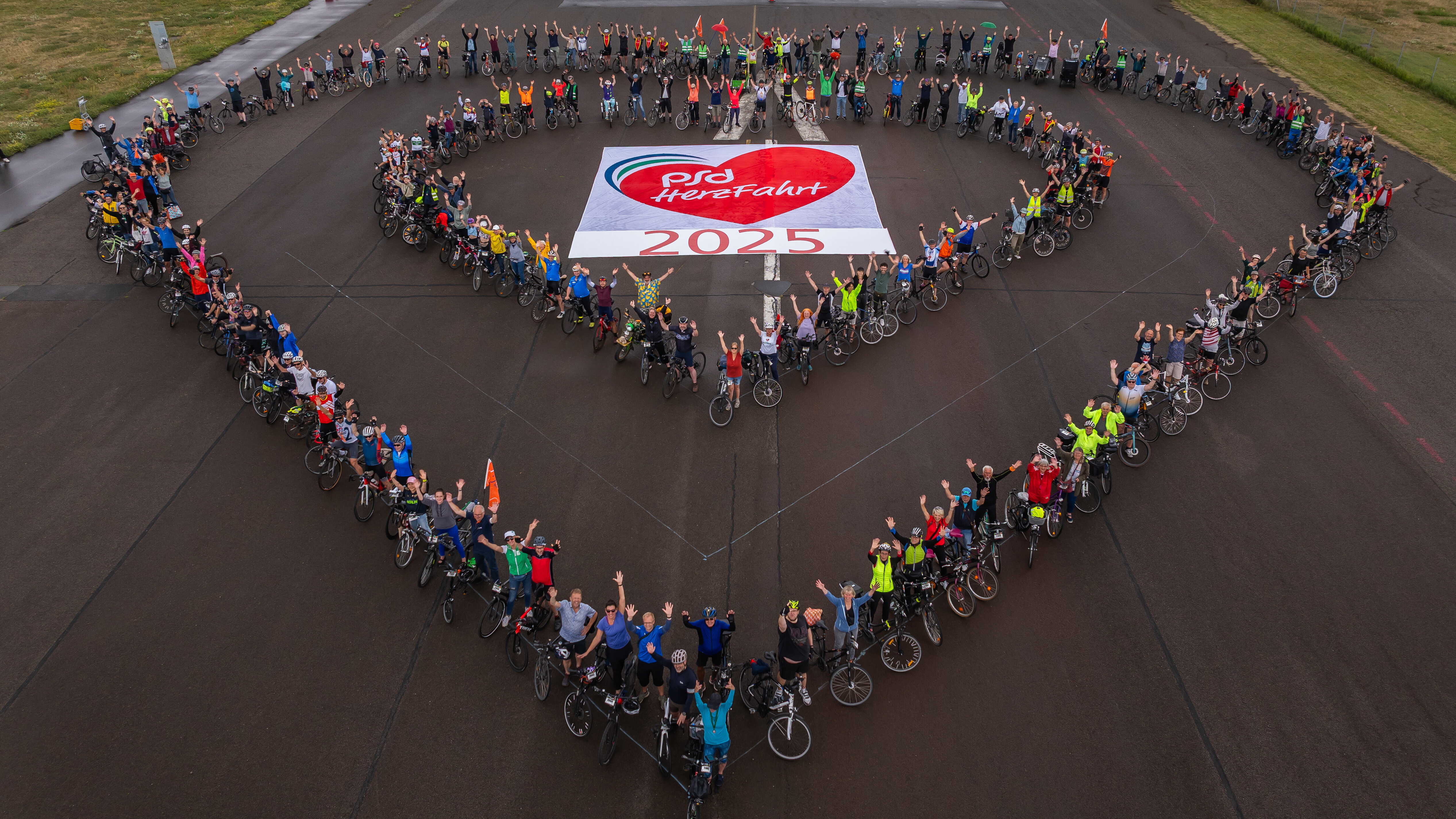 Eine Gruppe von Radfahrern steht auf einem Flugplatz und bildet gemeinsam ein Herz. In der Mitte liegt ein Banner mit der Aufschrift "PSG HereFahrt 2025". Viele Teilnehmer heben die Hände und sind in bunten Outfits gekleidet. Es herrscht eine fröhliche Atmosphäre.