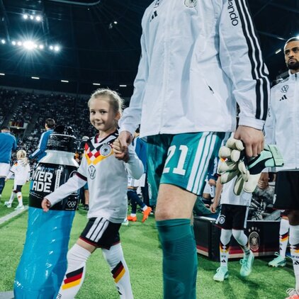 Ein deutscher Fußballspieler in einer weißen Jacke und grünen Sportshorts hält die Hand eines Mädchens, das ein Trikot der deutschen Nationalmannschaft trägt. Sie betreten gemeinsam das Spielfeld, umgeben von weiteren Spielern und einem Publikum.