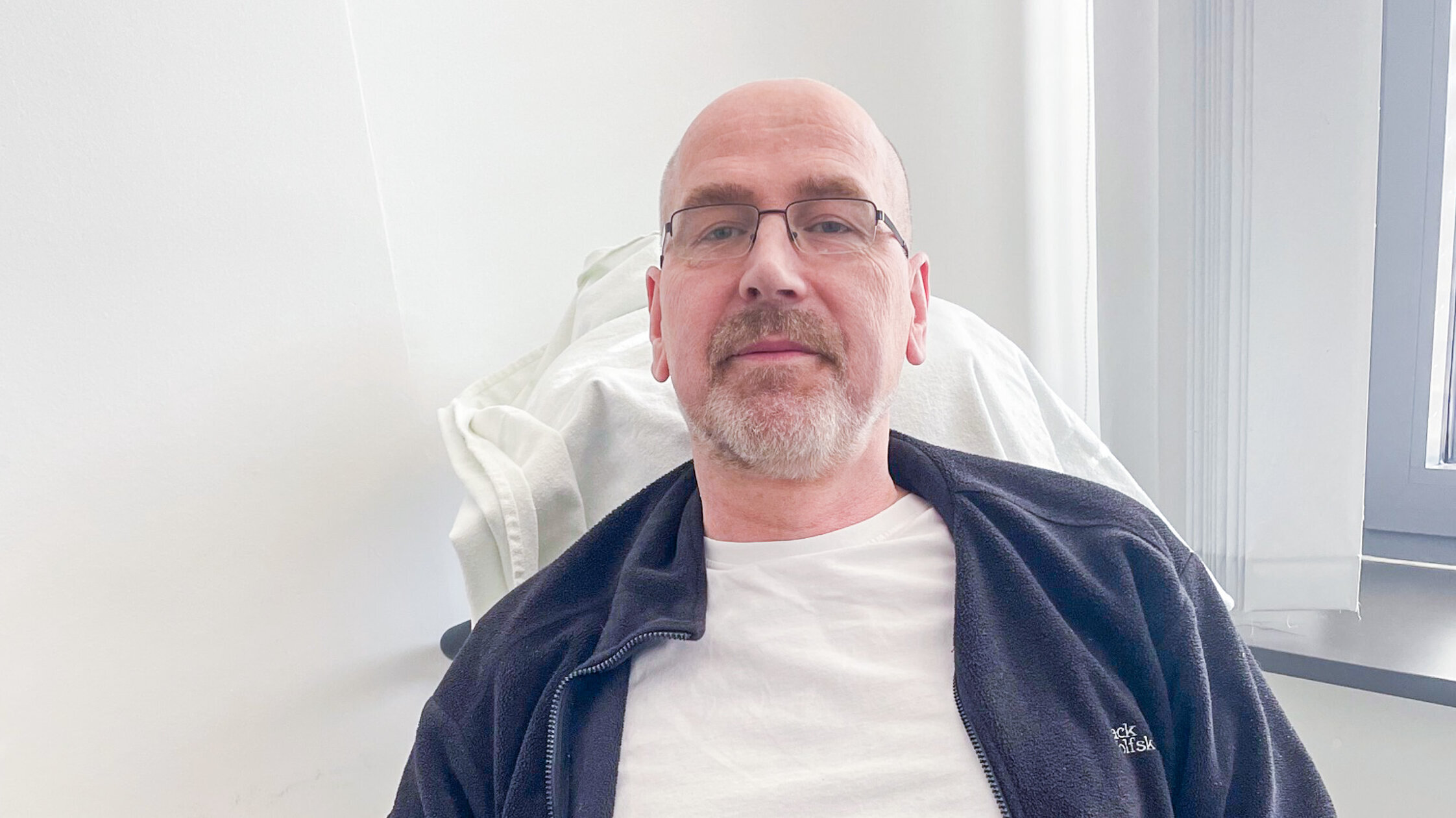 Ein Mann sitzt entspannt auf einem blauen Stuhl in einem hellen Raum. Er trägt ein weißes T-Shirt und eine schwarze Jacke. Der Mann hat eine Glatze und trägt eine Brille.