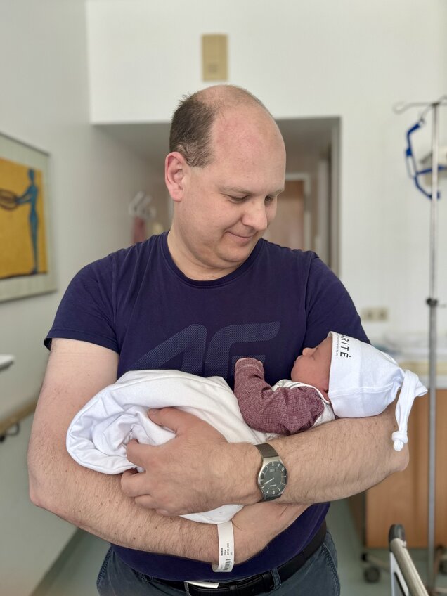 Ein Mann hält ein Neugeborenes in den Armen. Das Baby ist in eine Decke gewickelt und trägt eine Mütze. Der Mann schaut liebevoll auf das Baby, während sie in einem Krankenhauszimmer stehen.