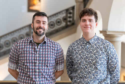 Zwei Männer stehen lächelnd nebeneinander auf einer Galerie. Der links trägt ein kariertes Hemd, der rechts ein gemustertes Hemd. Im Hintergrund sind architektonische Details und eine Wand mit Kunstwerken sichtbar.