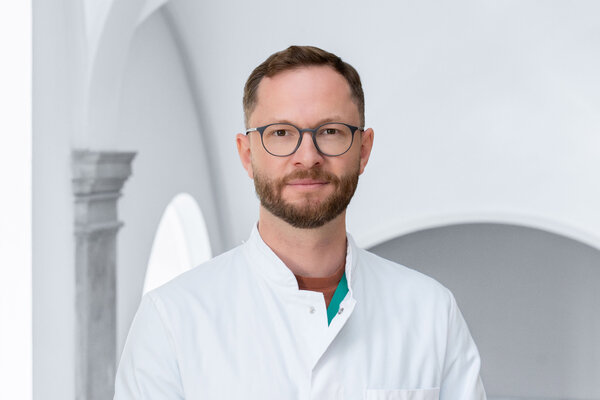 Ein lächelnder Mann mit Brille und einem weißen Arztkittel steht vor einem hellen, minimalistischen Hintergrund. Sein Gesicht drückt Freundlichkeit und Professionalität aus. Er hat einen gepflegten Bart und ist in einem professionellen Umfeld präsentiert.