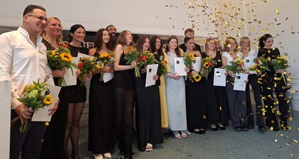 Eine Gruppe von Absolventen steht zusammen und hält Blumensträuße sowie Urkunden in den Händen. Über ihnen fliegen goldene Konfetti-Stücke. Die Szene strahlt Freude und Feierlichkeit aus, da sie einen wichtigen Meilenstein in ihrem Leben feiern.