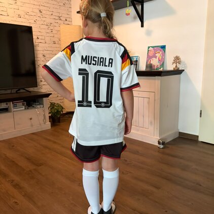 Ein Mädchen steht mit dem Rücken zur Kamera, trägt ein Fußballtrikot mit der Aufschrift "MUSIALA" und der Nummer 10. Es ist in den Farben der deutschen Nationalmannschaft gestaltet. Im Hintergrund sind ein Fernseher und einige dekorative Elemente zu sehen.