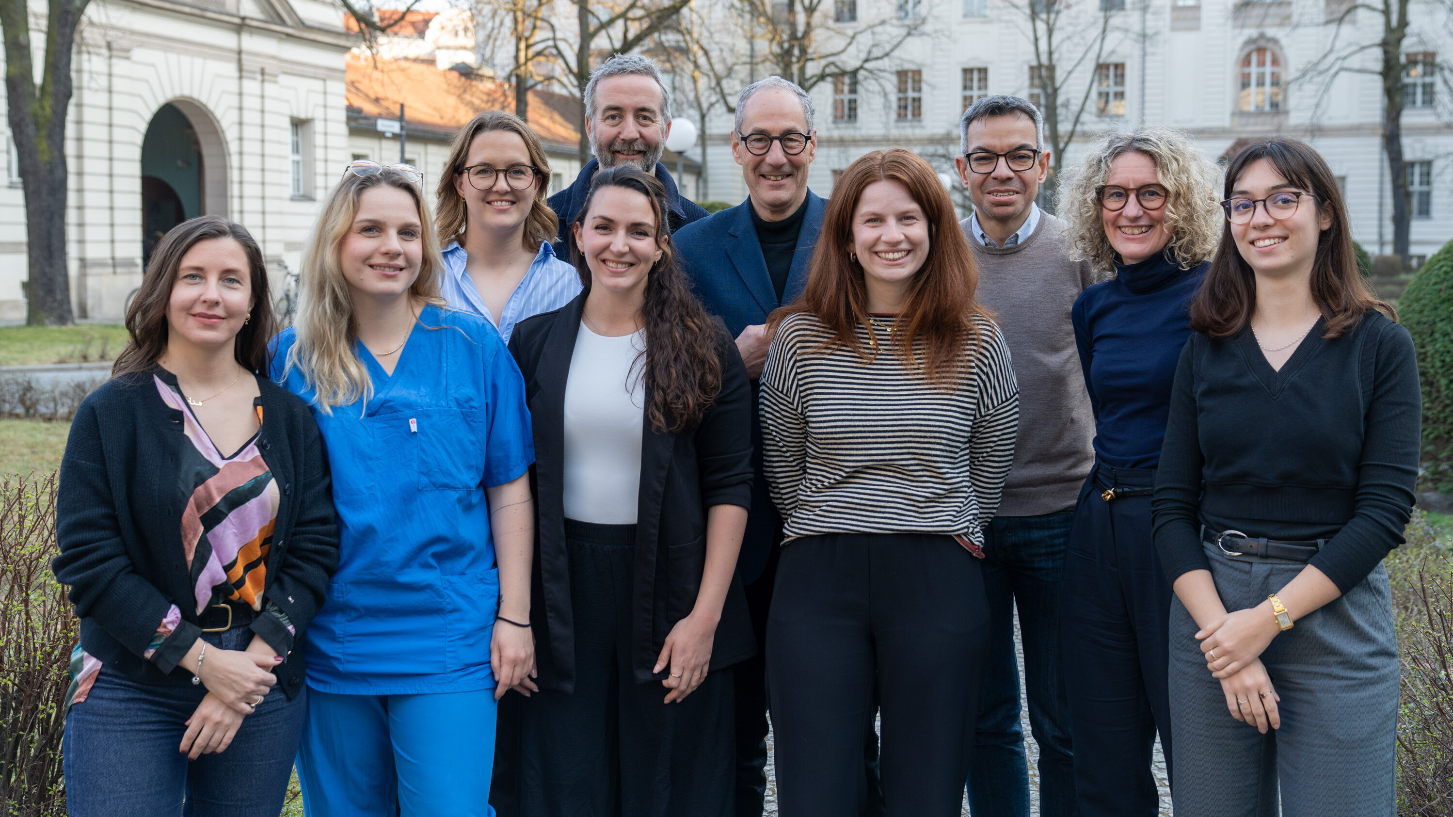 Eine Gruppe von zehn Personen steht im Freien, umgeben von Bäumen und einem Gebäude im Hintergrund. Die Gruppe setzt sich aus Männern und Frauen in verschiedenen Altersgruppen zusammen, die freundlich in die Kamera lächeln. Sie tragen unterschiedliche Kleidung, darunter medizinische und informelle Outfits.