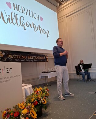Ein Mann steht auf einer Bühne und spricht willkommen, während eine Frau am Klavier spielt. Im Hintergrund ist ein Banner mit der Aufschrift „Prüfung bestanden! Wir sind stolz auf euch!“ und eine dekorative Blumenarrangement im Vordergrund.