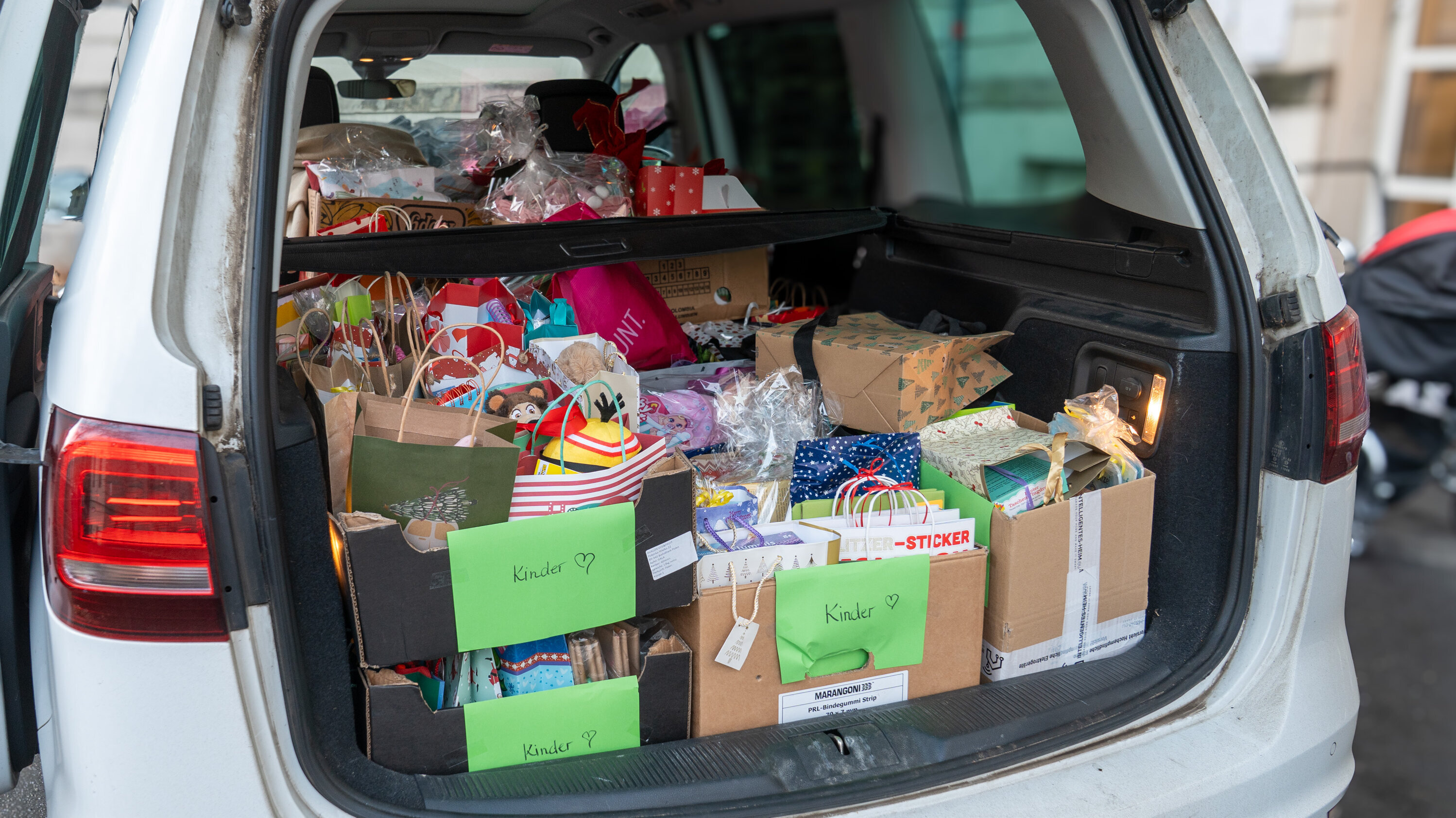 Ein offenes Auto mit einem Kofferraum voller Geschenkeboxen und Tüten, die farbenfrohe Verpackungen zeigen. Manche Boxen sind mit grünen Etiketten beschriftet, die das Wort „Kinder“ tragen. Die Geschenke sind sorgsam gepackt und scheinen für Kinder gedacht zu sein.