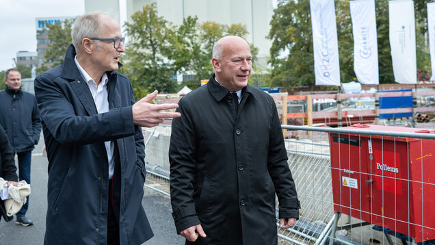Zwei Männer in formeller Kleidung unterhalten sich auf einer Baustelle. Der Mann auf der linken Seite gestikuliert mit der Hand, während der andere aufmerksam zuhört. Im Hintergrund sind Bauarbeiten erkennbar, umgeben von Bauzäunen und Maschinen.