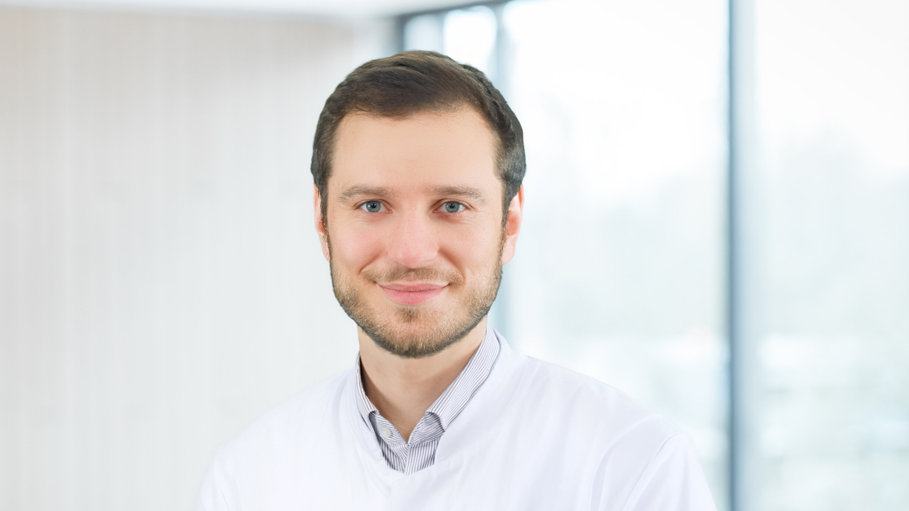 Ein junger Arzt in einem weißen Kittel lächelt freundlich in die Kamera. Im Hintergrund sind verschwommene Fenster, die Tageslicht hereinlassen. Sein Gesichtsausdruck wirkt sympathisch und einladend.