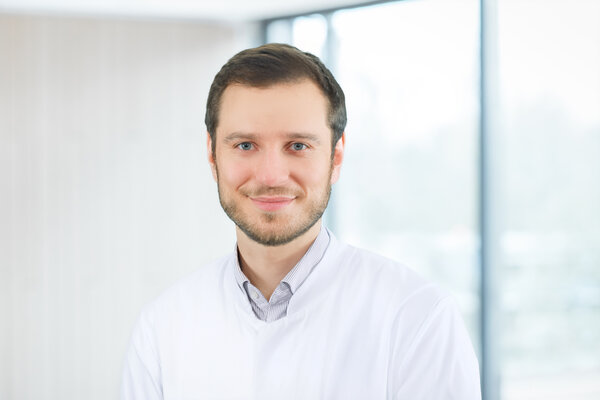 Ein junger Arzt in einem weißen Kittel lächelt freundlich in die Kamera. Im Hintergrund sind verschwommene Fenster, die Tageslicht hereinlassen. Sein Gesichtsausdruck wirkt sympathisch und einladend.